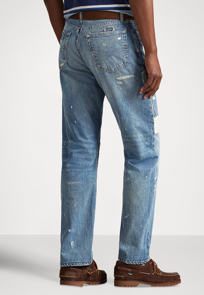 Uomo con jeans azzurri slavati con schizzi di vernice e scarpe casual in camoscio marrone, in piedi contro uno sfondo semplice.