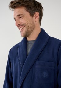 Navy-farbener Fleece-Bademantel mit Schalkragen, Fronttasche und dezentem Emblem. Weiche Textur und lockere Passform, ideal zum Entspannen.