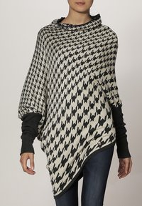 Zwarte en crème houndstooth poncho met een gedrapeerde vorm, voorzien van een hoge kraag en contrasterende zwarte sweatermouwen. Zachte textuur.