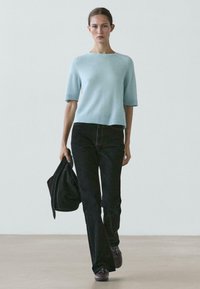 Maglione azzurro a maniche corte lavorato a maglia con scollo a coste, abbinato a jeans neri svasati e scarpe scure, completato da una giacca nera.