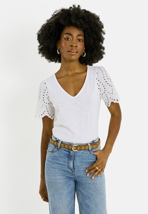 Mujer con cabello rizado que lleva una blusa blanca de escote en V con mangas abullonadas con ojales, vaqueros azules, un cinturón marrón, pendientes de aro dorados y anillos.