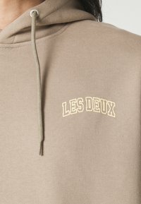 Felpa beige in tessuto morbido, con tasca a marsupio sul frontale e logo ricamato "LES DEUX" in giallo chiaro ad arco sopra il petto.