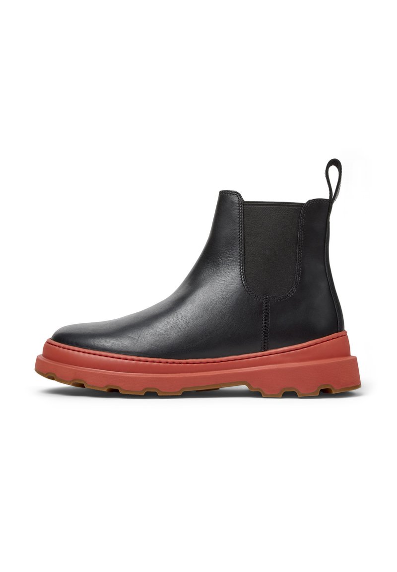 Musta nahkainen Chelsea-boot, jossa on oranssinpunainen kumipohja, varustettuna joustavilla sivupaneeleilla ja vetotarralla, esitettynä valkoista taustaa vasten.