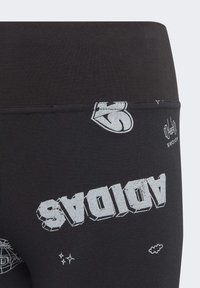 Leggings negros con impresiones gráficas plateadas, que incluyen "ADIDAS" en fuente negrita, motivos de estrellas y nubes, y una textura suave y elástica.