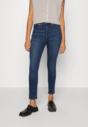Jeans skinny taille haute bleu foncé avec une texture lisse, des coutures discrètes et un ourlet au niveau de la cheville, accompagnés de chaussures noires à enfiler.
