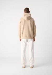 Beige hoodie with a drawstring hood, cuffed sleeves, and waistband, paired with white loose-fitting trousers and tan shoes.