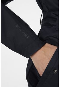 Manica di una giacca impermeabile nera con tessuto testurizzato, cuciture visibili e un logo del marchio impresso sul polsino.
