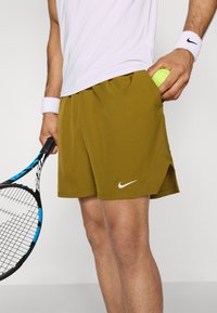 Nike Herren Sportshorts, olivgrün, leichtes Material, elastischer Bund, seitliche Taschen mit einem Tennisball und einem weißen Logoakzent.