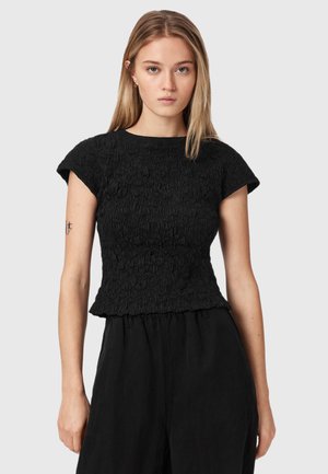 Jeune femme aux cheveux blonds et raides porte un haut noir texturé à manches courtes et un pantalon noir ample, debout devant un fond gris clair.