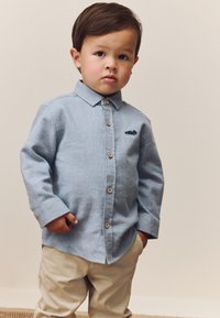 Camicia azzurro chiaro con bottoni, colletto, dettaglio ricamato di un'auto, bottoni in legno, abbinata a pantaloni beige. Tessuto dalla texture liscia.