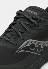 Mustad mustus Saucony jooksujalats, millel on mustrilised paelad, tekstureeritud põhi ja hõbedane logo küljes, lähedal vaatamine ülemisele esiosale.