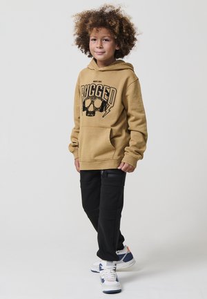 Niño con cabello rizado que lleva una sudadera color beige con el texto "RUGGED" y un gráfico de calavera, pantalones negros y zapatillas blancas, de pie frente a un fondo liso.