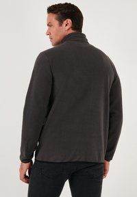 Mørkegrå fleecejakke med høj krave, glat tekstur og lange ærmer. Bagfra ses et enkelt design uden synlige lukninger.