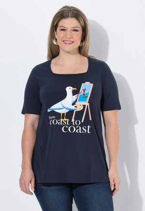 Smilende kvinde iført en mørkeblå skjorte med en maleri af en måge på et staffeli og teksten "fra kyst til kyst."