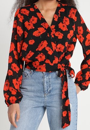 Kvinde iført sort omslagsbluse med rødt blomsterprint og lyseblå jeans med høj talje, med bindedetalje i taljen på blusen.