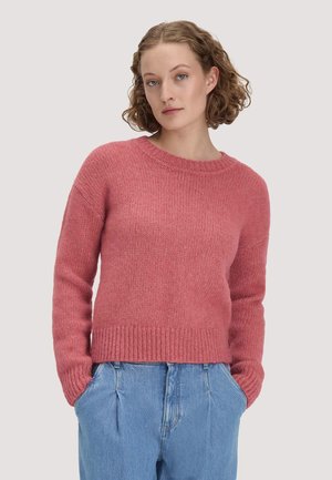 Femme aux cheveux courts et bouclés portant un pull en maille rose et un jean bleu taille haute, les mains dans les poches, sur un fond uni.