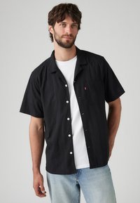 Zwart shirt met korte mouwen met een textuurpatroon, witte knopen en een borstzak; gedragen over een wit T-shirt.