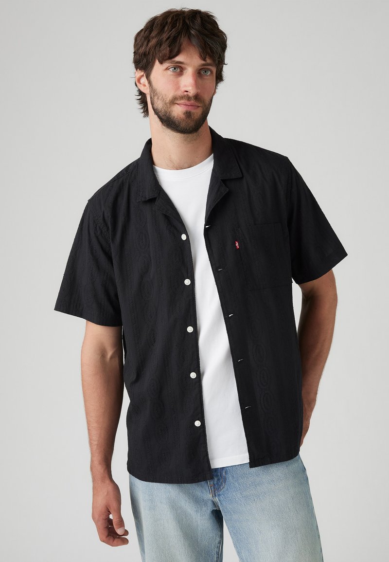 Zwart shirt met korte mouwen met een textuurpatroon, witte knopen en een borstzak; gedragen over een wit T-shirt.