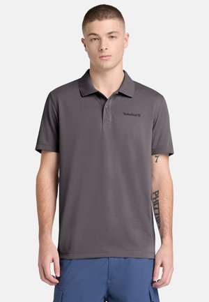 Junger Mann mit kurzen Haaren, der ein dunkelgraues Timberland-Poloshirt und blaue Cargo-Shorts trägt, vor einem hellen Hintergrund stehend.