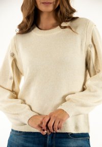 Suéter color crema con puños acanalados y cuello redondo. Presenta botones decorativos a lo largo de las mangas. Tela suave y texturizada.