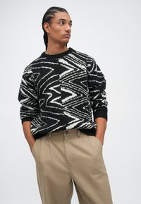 Junger Mann mit geflochtenem Haar, der einen schwarz-weißen Zickzackmuster-Pullover und beigefarbene Hosen trägt, mit den Händen in den Taschen.