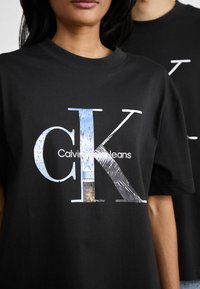 Crna pamučna majica s velikim grafičkim prikazom s tekstom "CK" i "Calvin Klein Jeans" u bijeloj boji te stiliziranim slikama unutar slova.