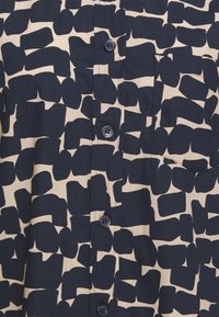 Camicetta a maniche corte con bottoni di colore beige con grandi motivi astratti blu navy. Presenta una tasca sul petto e bottoni rotondi blu navy. Tessuto di consistenza liscia.