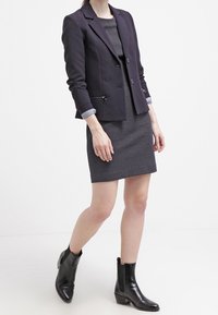 Granatowa marynarka z wyłogami i dwoma guzikami, zestawiona z teksturowaną szaro-sukienką. Czarne botki za kostkę z niskim obcasem dopełniają całość.