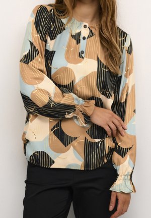 Femme portant un chemisier à manches longues avec un imprimé géométrique abstrait beige, noir et bleu clair, et un pantalon noir, une main touchant le bras opposé.