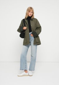 Olive zielona, oversize kurtka z bocznymi kieszeniami, zestawiona z czarnym golfem, jasnoniebieskimi spodniami w szerokim kroju i białymi trampkami.