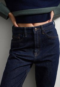 Jeans de mezclilla azul oscuro con costuras en contraste, que cuentan con un cierre de botón y un diseño clásico de cinco bolsillos. Textura suave y forma ajustada.