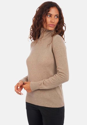 Femme aux cheveux bouclés portant un pull col roulé beige et un pantalon noir, regardant vers sa droite avec les mains jointes devant elle.