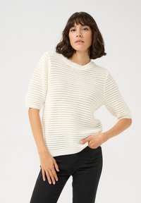 Pull en tricot blanc à manches courtes, avec un design texturé en ouverture. Associé à un pantalon noir avec une coupe décontractée.