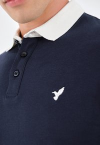 Marineblaues Polohemd aus Baumwolle mit weißem Kragen. Drei-Knopf-Leiste, gesticktes Vogel-Logo auf der linken Brust, glatte Textur.