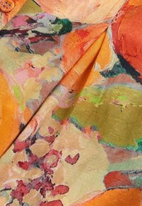 Farebná textília s abstraktnými kvetinovými vzormi v oranžovej, zelenej a ružovej farbe. Obsahuje detail gombíka a textúrovaný povrch.