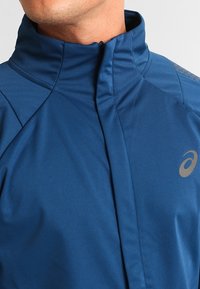 Uomo che indossa una giacca blu con zip, colletto alto e logo riflettente sul petto, contro uno sfondo bianco.