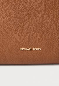 Καφέ δερμάτινη υφή με τελείωμα από μικρές κοκκώδεις λεπτομέρειες· διαθέτει χρυσή φίρμα "MICHAEL KORS". Απλός, κομψός σχεδιασμός με ορθογώνιο σχήμα.