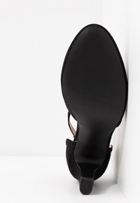 Chaussure noire à talons hauts vue de la semelle, montrant un bout pointu, une semelle inférieure à texture lisse et un talon étroit sur fond blanc.