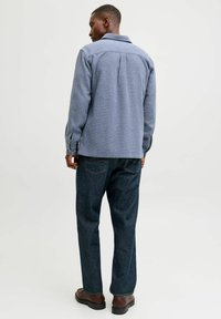 Blå teksturert skjorte med krage, synlige sømmer og knappede mansjetter, kombinert med mørke denimjeans og brune sko. Sett bakfra.