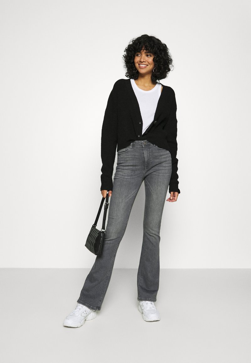 Schwarze Strickjacke, weißes T-Shirt, graue Schlagjeans und weiße Sneaker. Schwarze strukturierte Handtasche mit kurzem Tragegurt. Lockige Haare.