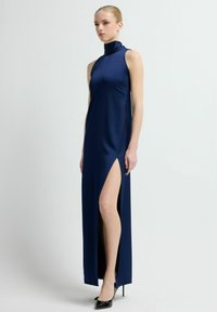 Granatowa, bezrękawnikowa sukienka maxi z wysokim kołnierzem i bocznym rozcięciem. Gładka tkanina o eleganckiej sylwetce, zestawiona z czarnymi szpilkami.