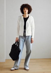 Bijela zip-up dukserica preko crne majice, uparena sa svijetloplavim širokim trapericama. Crna torba se drži u jednoj ruci. Neutralna pozadina.
