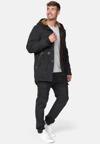 Chaqueta negra con capucha, forrada en fleece color beige y detalles marrones, combinada con pantalones cargo negros y zapatillas blancas.