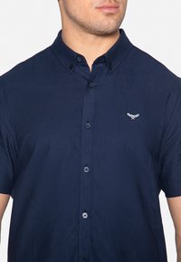 Marineblaues Kurzarmhemd mit Button-Down-Kragen, das ein dezentes gesticktes Logo auf der linken Brust und runde schwarze Knöpfe aufweist.