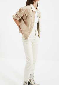 Beige džínsová bunda s bílým, strukturovaným límcem z beránka, v kombinaci s krémovými džíny a vzorovanými kotníkovými botami. Světlé pozadí.