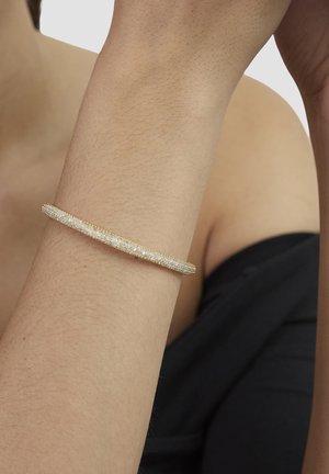 Gold-tone bangle bracelet with a textured surface, slim design, and subtle sparkle, worn on a brown arm against a dark background.