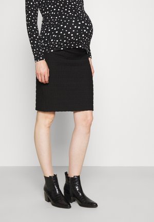 Zwangere vrouw die een zwart polka dot top draagt, een zwarte, textuurrijke rok tot op de knie en zwarte enkellaarsjes met hak, staand tegen een effen achtergrond.