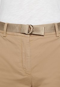 Ceinture en tissu beige avec boucle double anneau métallique portée sur un pantalon beige et une chemise blanche. La ceinture comporte un texte brodé subtil.