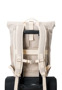 Beige hátizsák párnázott pántokkal, amely egy fekete bőrönd fogantyújához van rögzítve, hátulról, fehér háttér előtt.