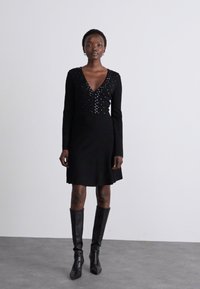 Abito nero con maniche lunghe, scollatura a V profonda, colletto decorato con elementi brillanti e corsetto aderente che si trasforma in una gonna svasata.